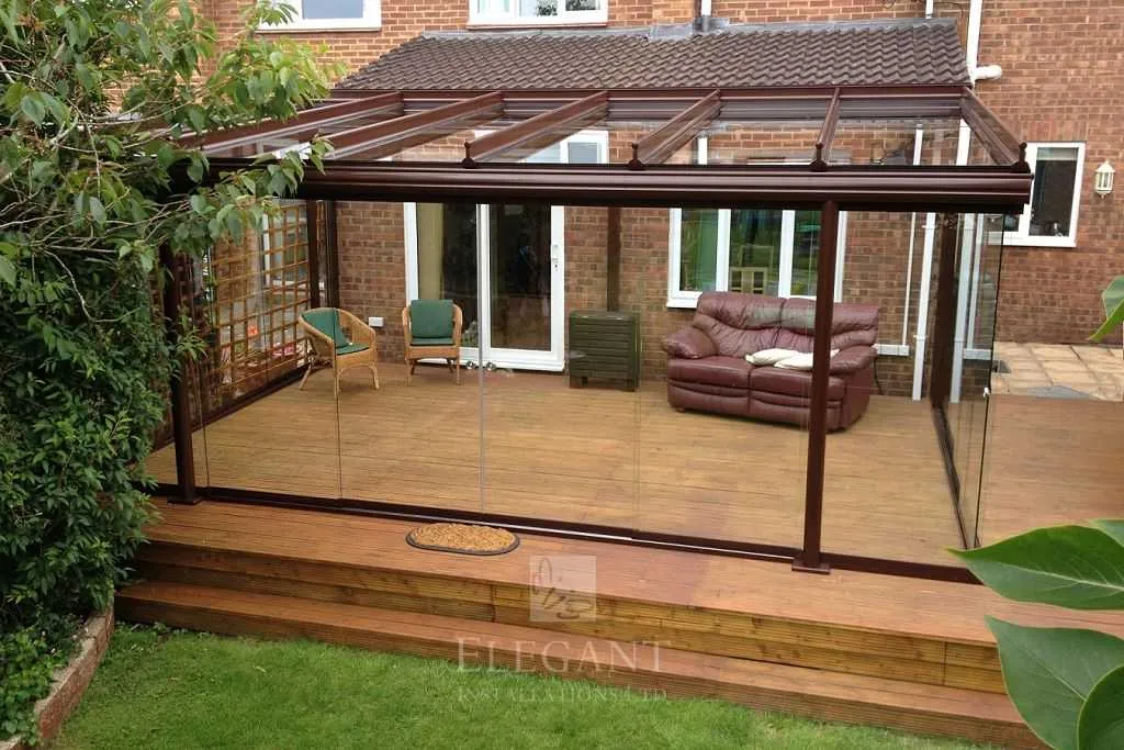 Modern glass veranda extension with wooden decking and outdoor seating attached to a brick house.